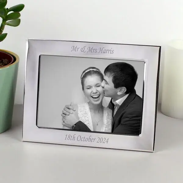 Silver photo frame engraved with Mr and Mrs Harris and a date, displaying a black and white picture of a couple embracing, placed on a surface beside a plant and candle.